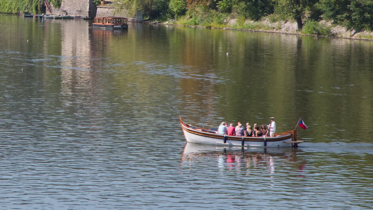 Small passenger boat with tourists moves gently on calm river, sunny day, steady wide shot