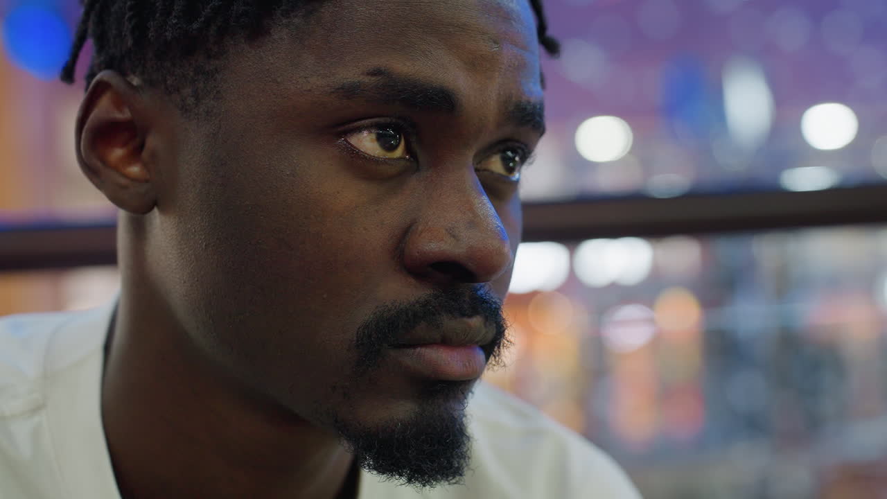 Close up of man with serious contemplative look seated in mall as he checks his time with blurred bokeh lights in background creating thoughtful mood of waiting and patience indoors