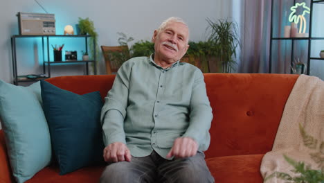 grandfather man smiling friendly at camera waving hands gesturing hello hi greeting at home on sofa