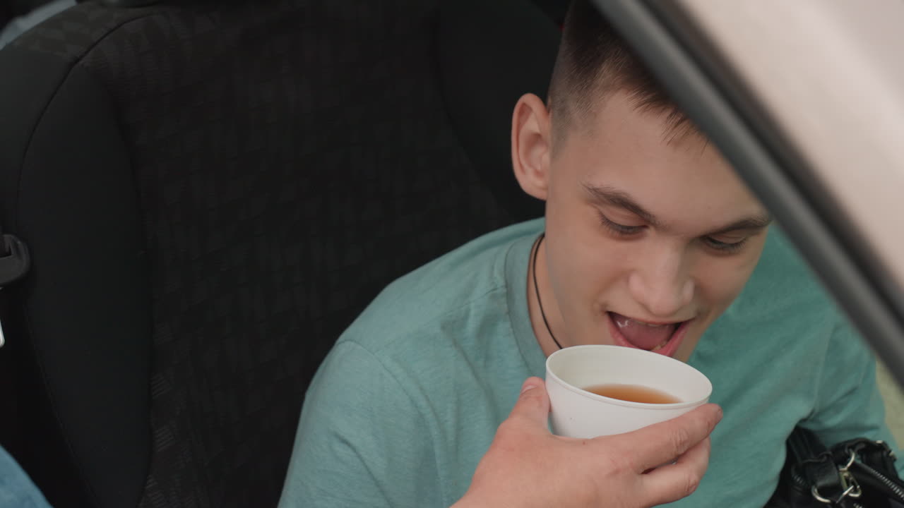 White Young Man Tasting Warm Drink From Cup In Car Seat With Support Worker Candid Closeups Capture Hopeful Expression, Assisted Sip, Wheelchair Access And Companion Care, Quiet Transport Scene That