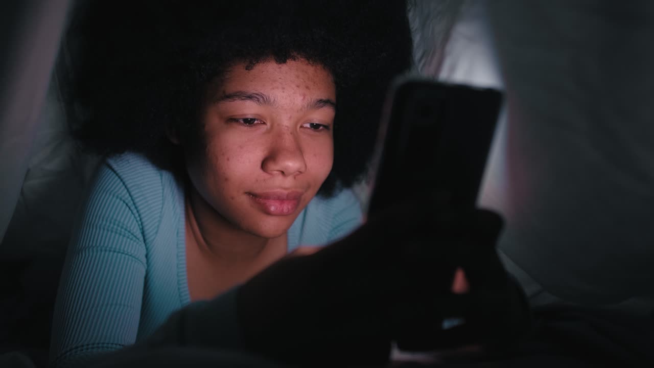 Cheerful teenage girl browsing mobile phone in the living room at night