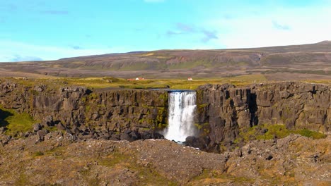 Aerial-drone-shot-above-a-large-Icelandic-plain-with-a-large-waterfall-flowing-through-a-fault-on-land,-sunny-weather-and-blue-sky