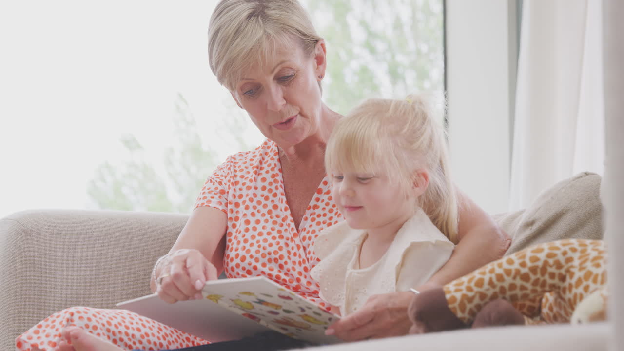 집에서 손녀와 함께 소파에 앉아서 책을 읽고 있는 할머니
