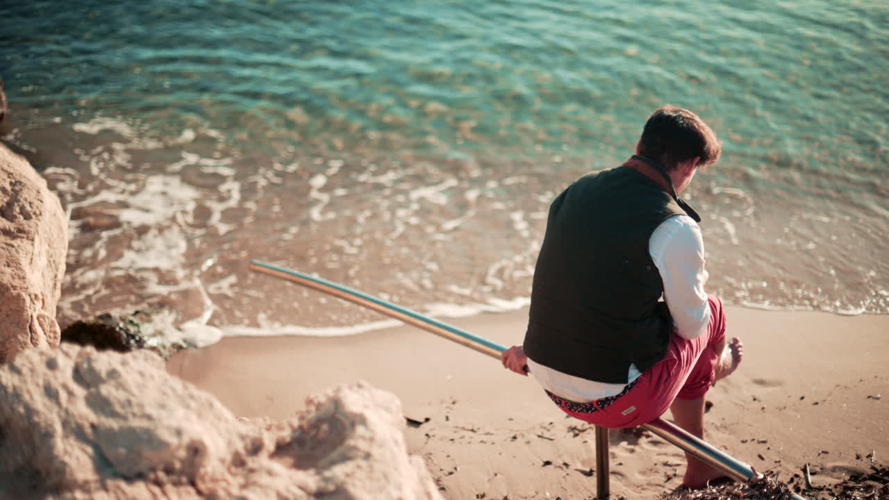 A young man sits on a metal rail at the edge of a sandy beach, looking toward the calm sea in warm afternoon light