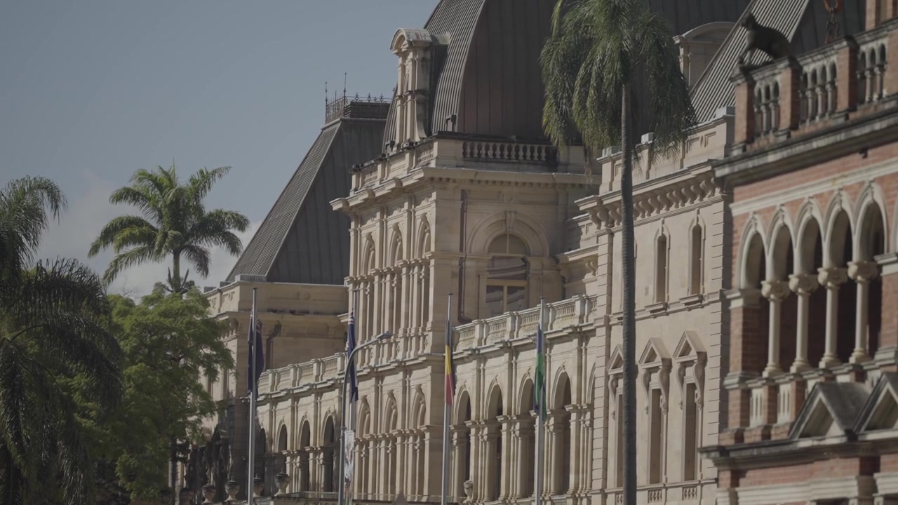Une rue bordée d'arbres longe un bâtiment patrimonial orné avec des drapeaux flottant à son entrée à Brisbane