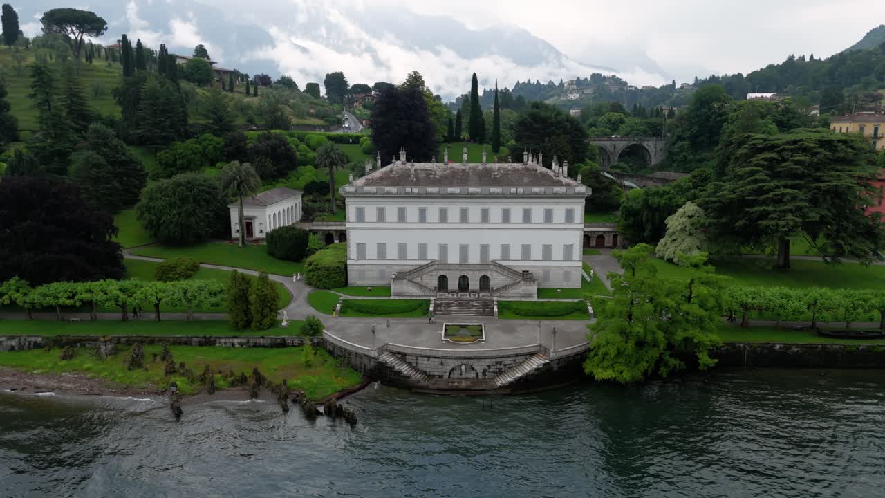 Flying backwards from the famous luxury wedding Venue Villa Melzi d´Eril in Bellagio italy at lake como in its neiclassical architecture revealing the nature and alp mountains covered in clouds