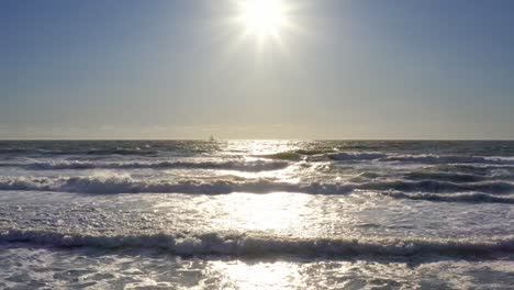 Drone-4K-low-altitude-push-over-beach-waves-toward-sunset-with-sailboat-in-distance-and-birds