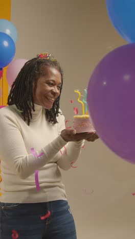 vertical video studio shot of woman wearing birthday headband celebrating birthday blowing out candles on cake with paper confetti