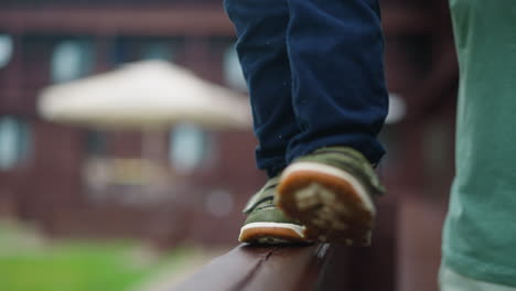 Toddler-boy-in-green-sneakers-walks-along-wooden-railing