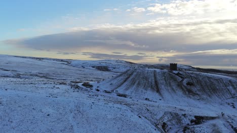 Snowy-Rivington-Pike-tower-Winter-hill-aerial-view-people-sledding-downhill-at-sunrise-right-pan