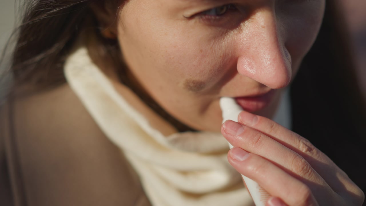 Primo piano di donna che applica lucidalabbra con mano ferma, concentrandosi sul processo di applicazione. Il soggetto indossa una sciarpa chiara e un capospalla marrone; l'illuminazione soffusa evidenzia i dettagli della texture della pelle.