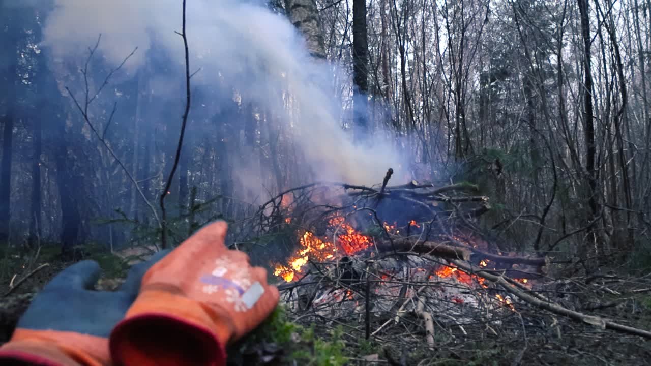 Low angle footage of a bonfire or a camp fire burning in a forest with thick white and blue smoke coming out from top in slow motion while some working protective gear gloves in front during winter.
