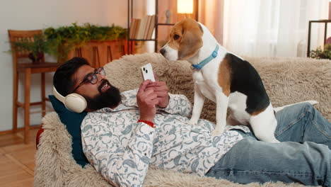 indian man lies on sofa listening to music or audiobook while beagle dog calmly rests on stomach