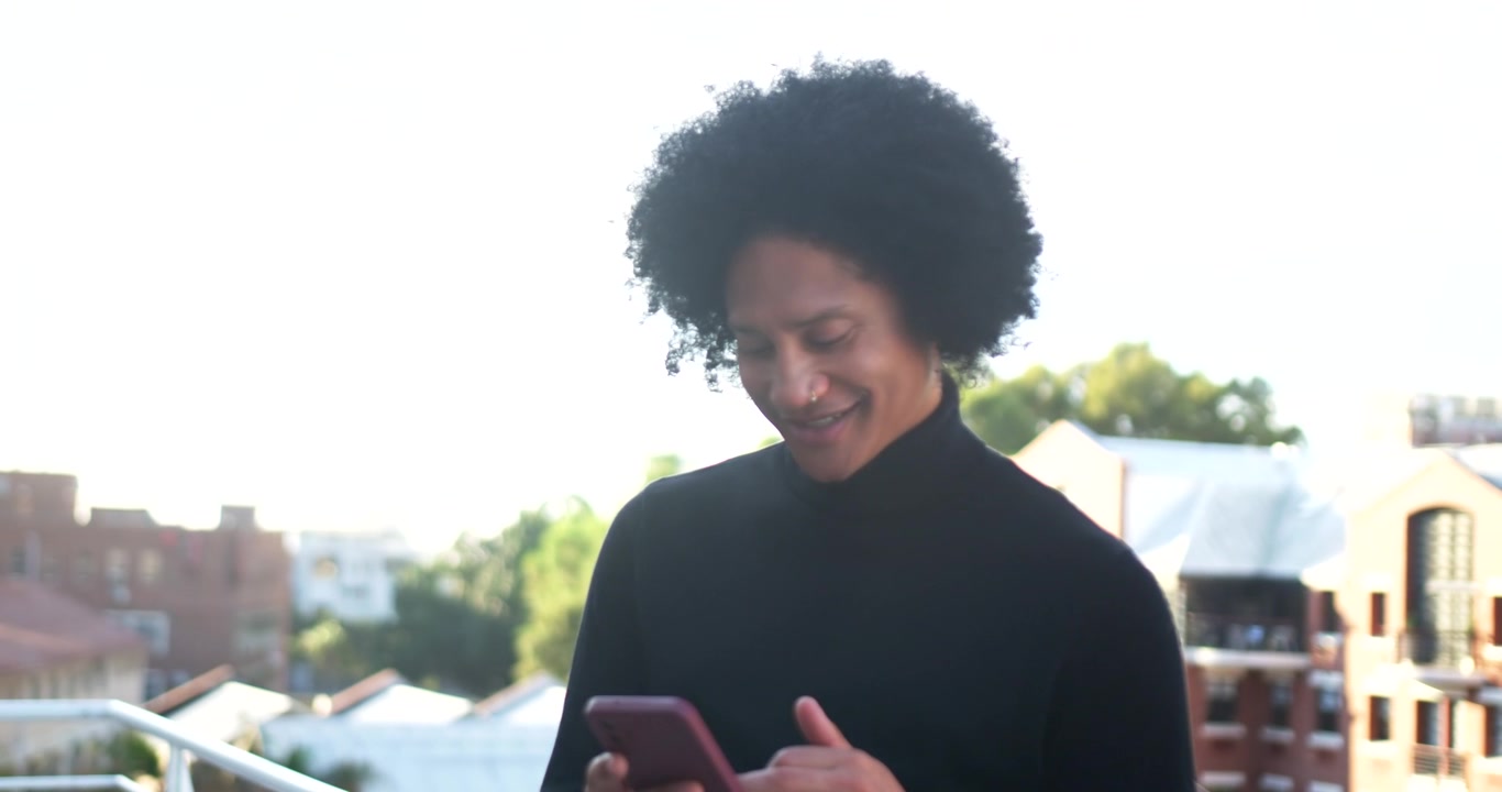 Uomo afroamericano che solleva lo smartphone, toccando, scorrendo e leggendo messaggi su un balcone sul tetto