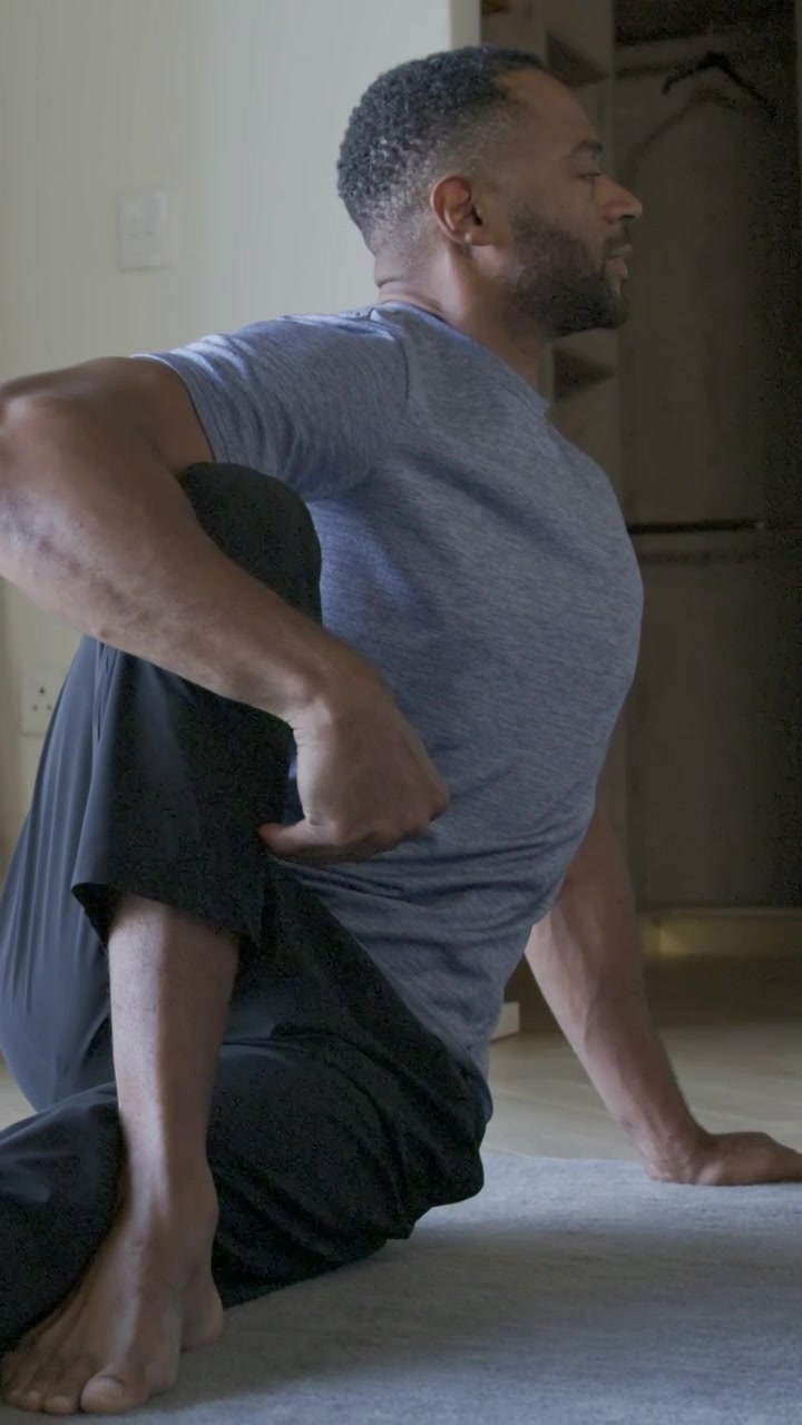 Vertical video: Starting stretch, man in gray tee doing spinal twist on home carpet holding knee