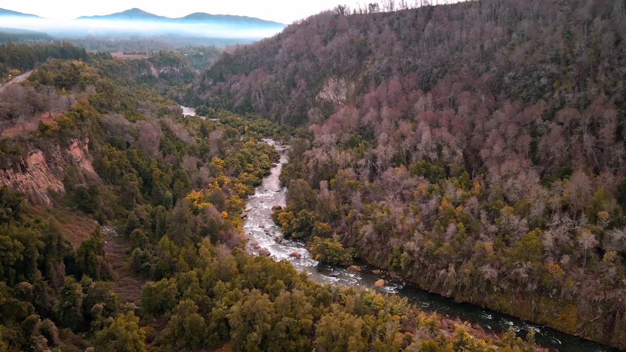 Orbiting drone aerial of turquoise river through misty canyon valley with autumn forest, dramatic mountains and fog backdrop, Chile wilderness