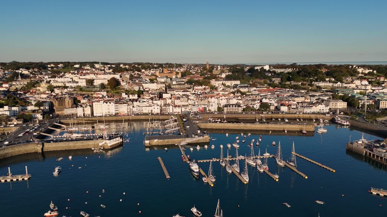 Metraje de dron de gran altura que muestra el puerto de St Peter Port en Guernsey, sus amarres de pontones exteriores, la piscina y vistas extensas de St Peter Port bajo el sol de la tarde en un día despejado.