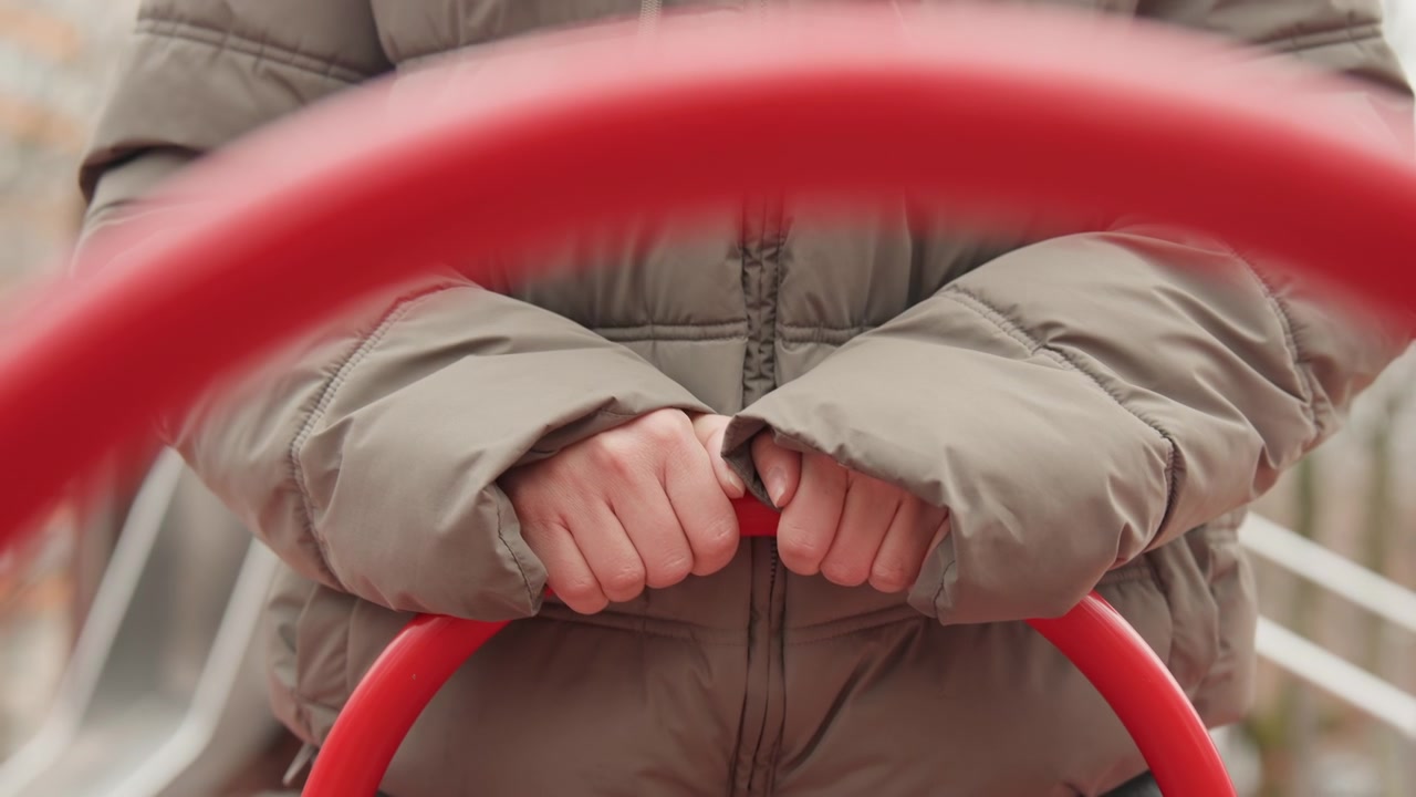 Primo piano di mani che afferrano una sbarra rossa in un parco giochi vuoto, persona con giacca a piumino color oliva che tiene un anello di metallo, luce invernale attenuata, profondità di campo ridotta, bokeh morbido, immobilità contemplativa, nocche in evidenza.