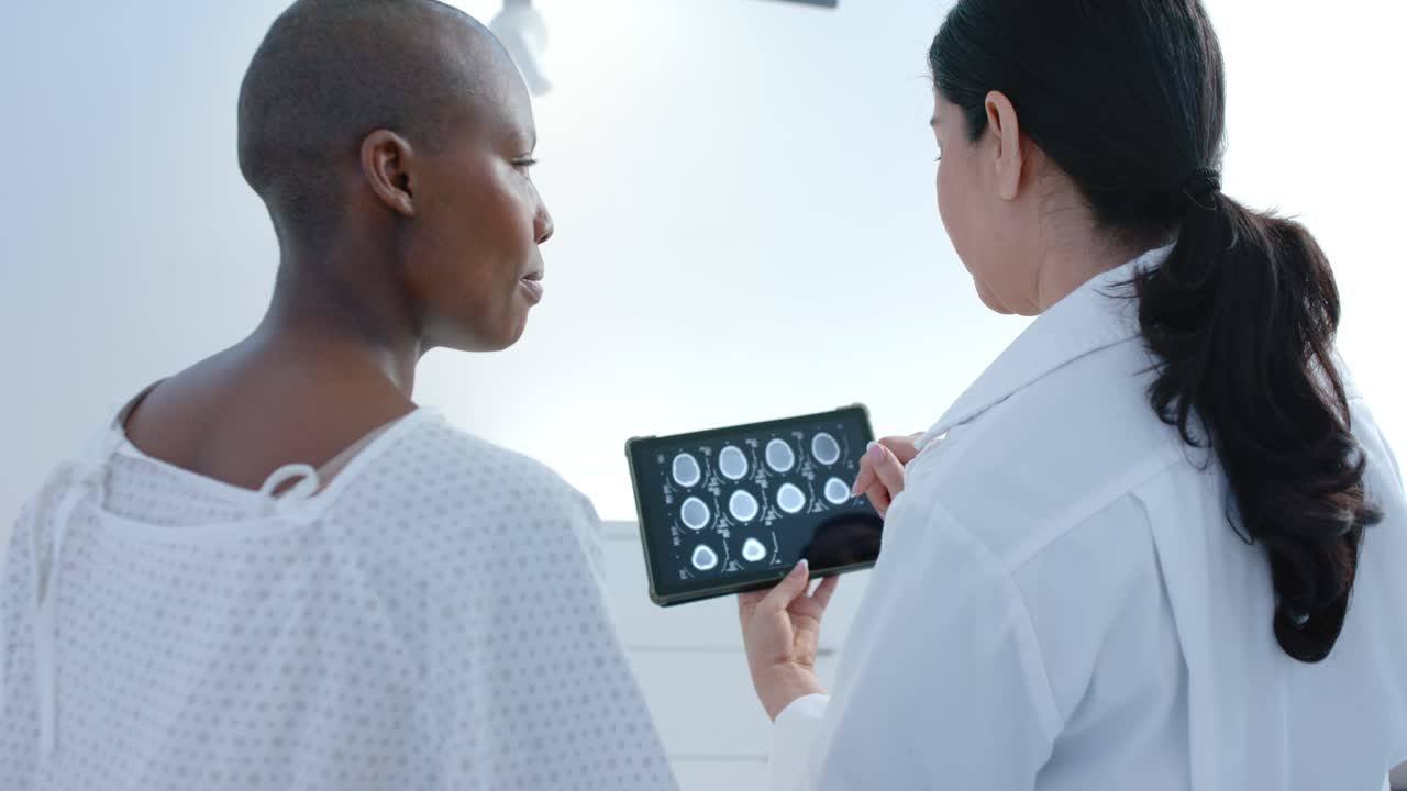 Dotteressa e paziente di etnie diverse, entrambe in camice, che visualizzano scansioni su un tablet in una clinica, con la dotteressa che tocca lo schermo e spiega i dati.