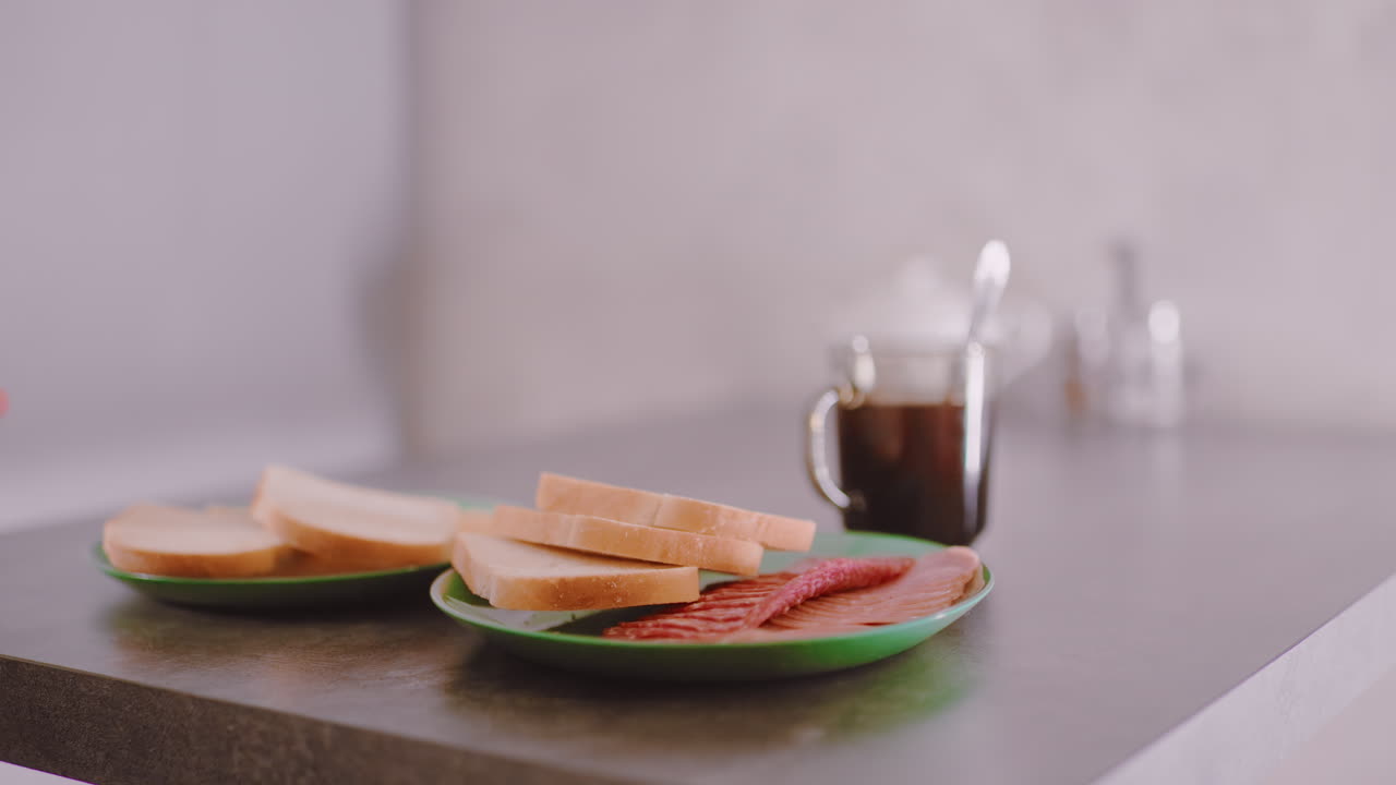 Hand holding green plate with bread and meat moving toward table near hot cup of coffee, illustrating simple home dining preparation with warm domestic atmosphere