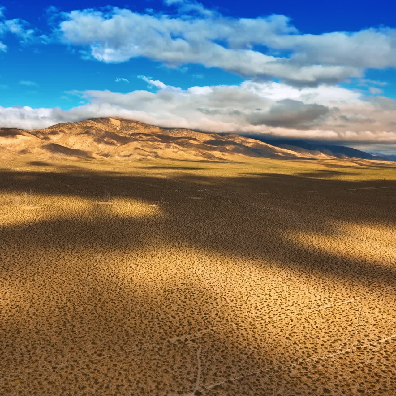 Dry deserted land with some spots of desert plants. Clouds float by the sky and throw shadows on the landscape. Top view