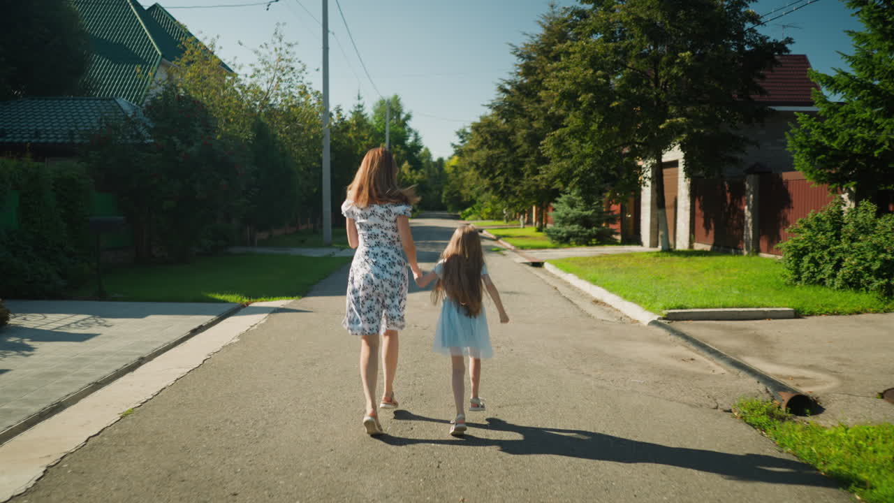 Rear view of mother and daughter holding hands while running together along peaceful residential street in daylight, floral dresses flowing as sunlight