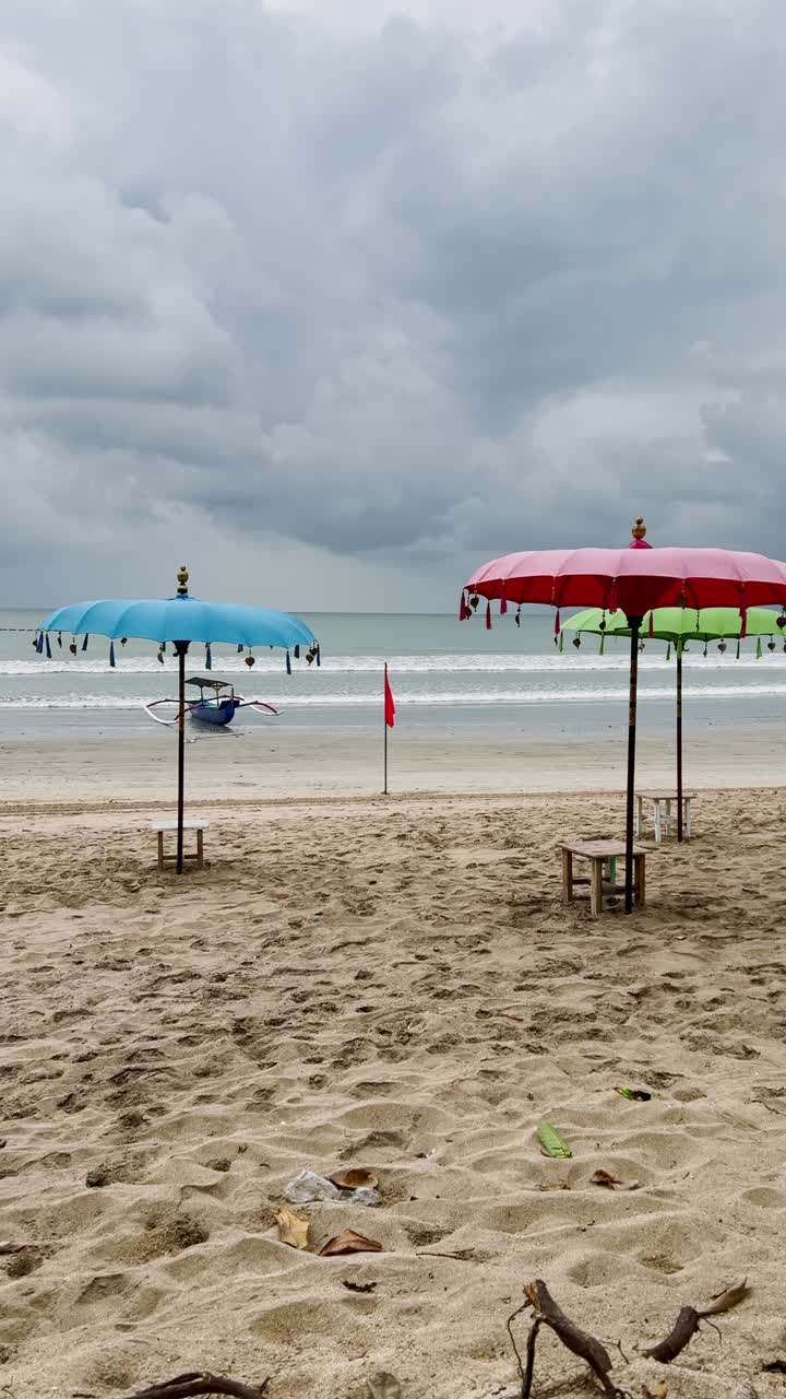 Vertical video capturing colorful umbrellas along Kuta Beach Bali with ocean waves and tropical charm under dramatic storm clouds, creating a coastal scene rich in atmosphere and island beauty