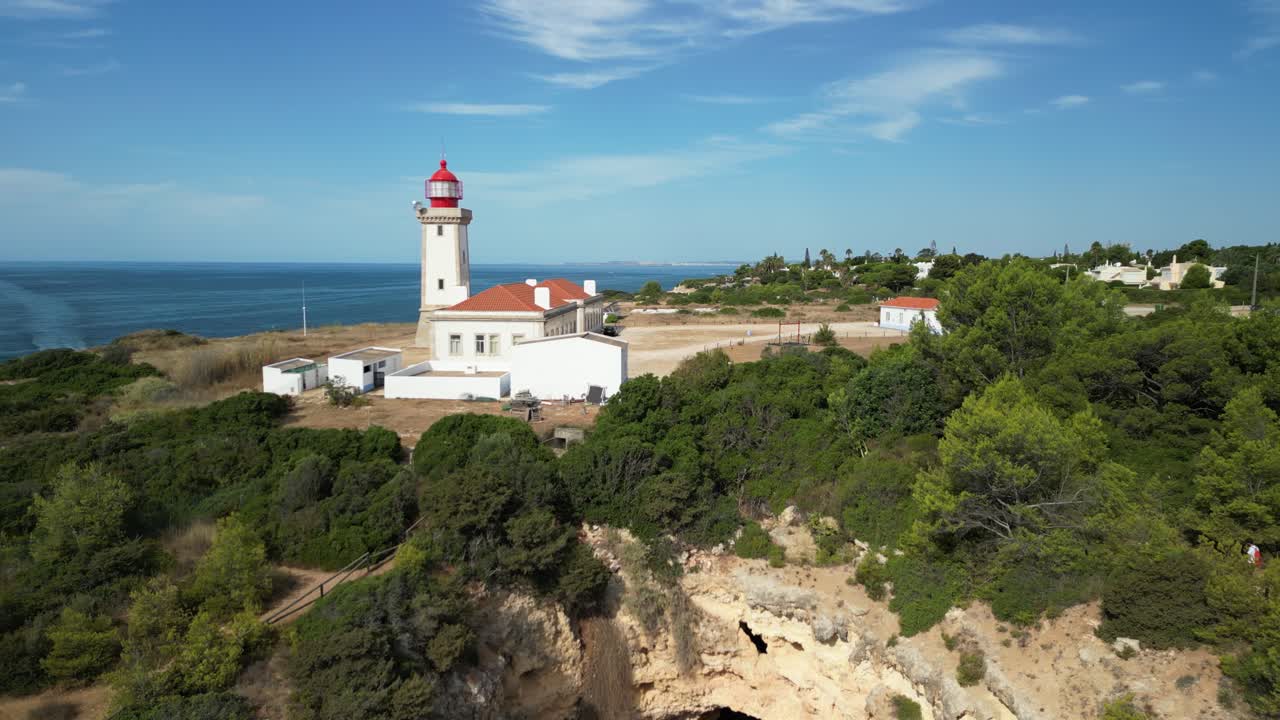 Drone shot of red roofed lighthouse near rocky coastline in Faro, Portugal, surrounded by green trees, clear sky, calm blue ocean, coastal cliffs, scenic view, peaceful atmosphere, summer day