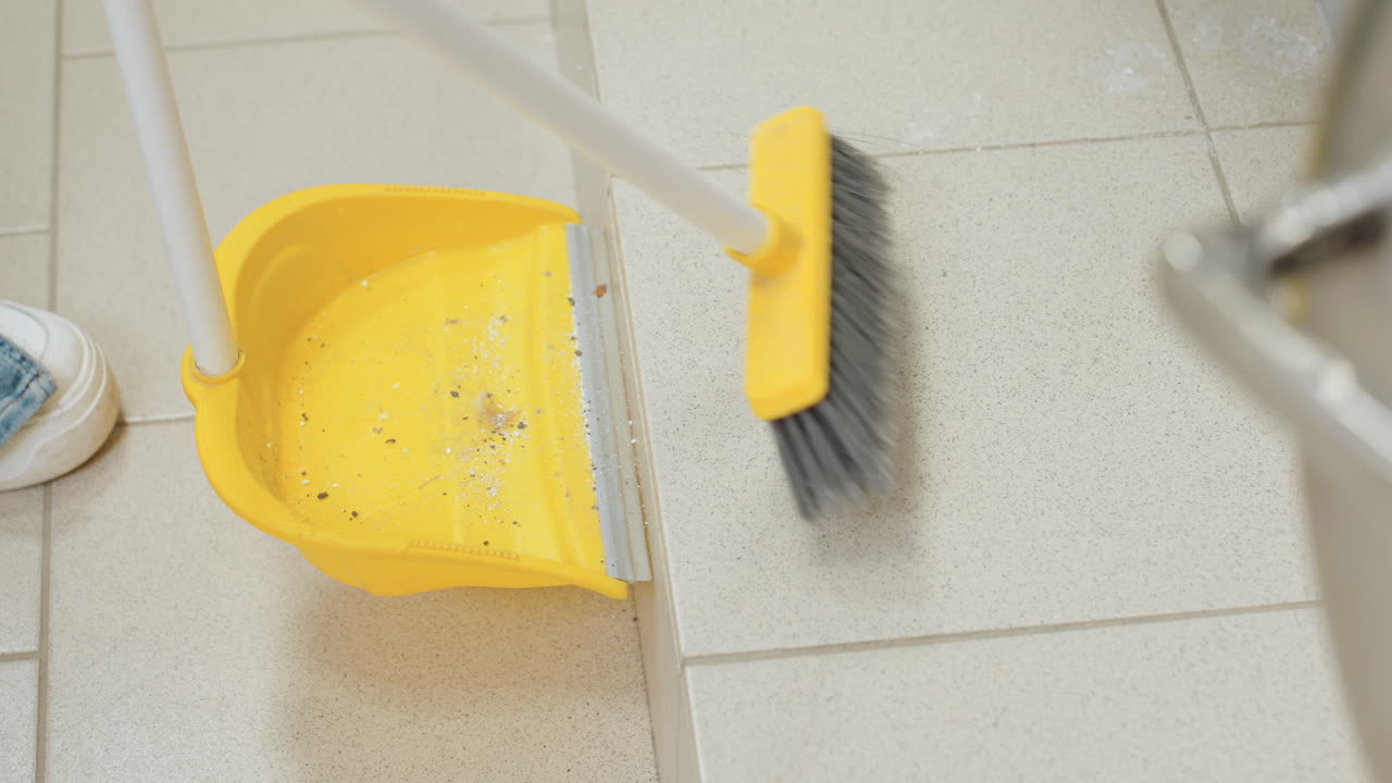 High angle view shows hand sweeping dirt into dustpan with broom on tiled floor beside stainless washer, careful cleanup, hygiene, sanitation, bristles push debris toward pan during maintenance