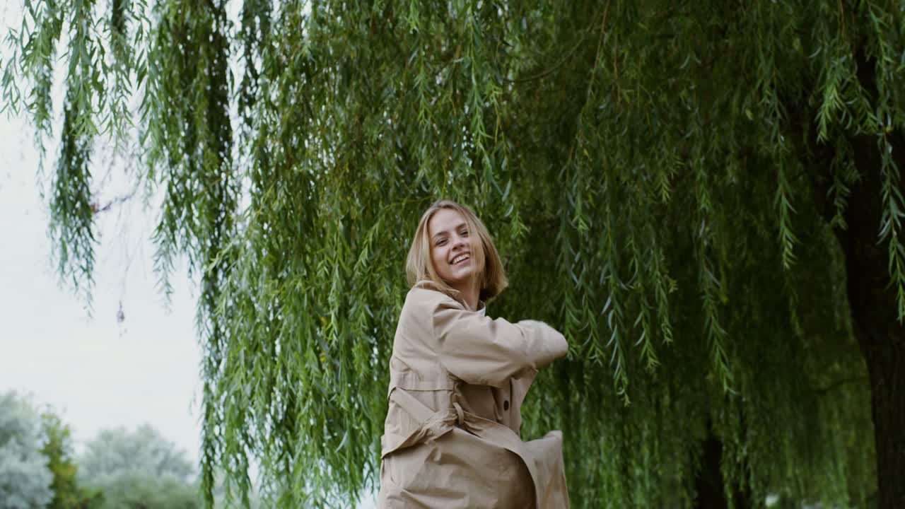 Happy Woman in a Beige Trench Coat under a Willow Tree