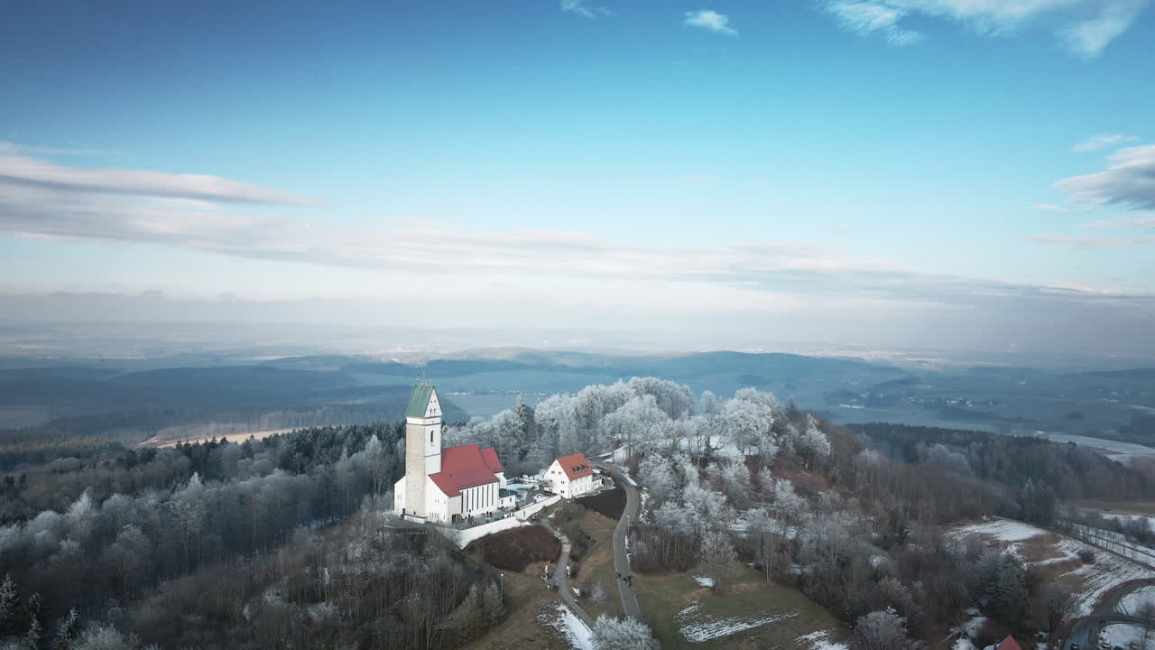 Un drone esegue una fluida orbita in senso orario attorno alla prominente cappella bianca sulla collina di Utterweiler, Baden-Württemberg, mostrando vaste foreste ghiacciate, prati innevati e una valle lontana.