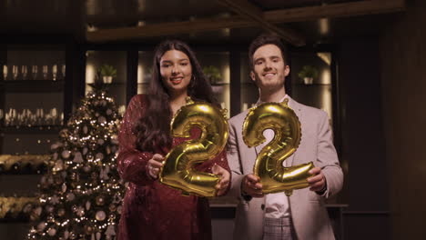 woman and man wearing elegant clothes holding balloons with the numbers 22 while dancing, kissing and hugging each other in new year's party