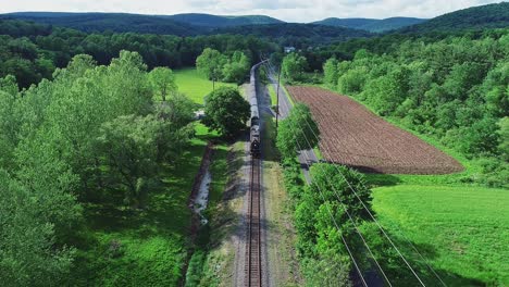 an aerial view of a long steam passenger train approaching on a single track traveling thru green farmlands on a spring day