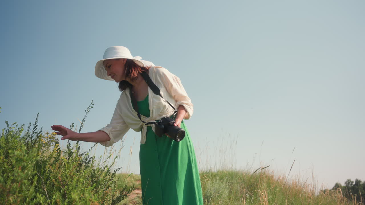 Photographer in green dress and wide hat gently touches yellow wildflower while walking through lush grassy hill holding camera, enjoying serene summer scenery under clear blue sky