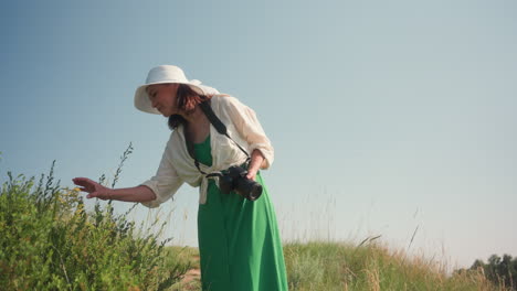photographer in green dress and wide hat gently touches yellow wildflower while walking through lush grassy hill holding camera, enjoying serene summer scenery under clear blue sky
