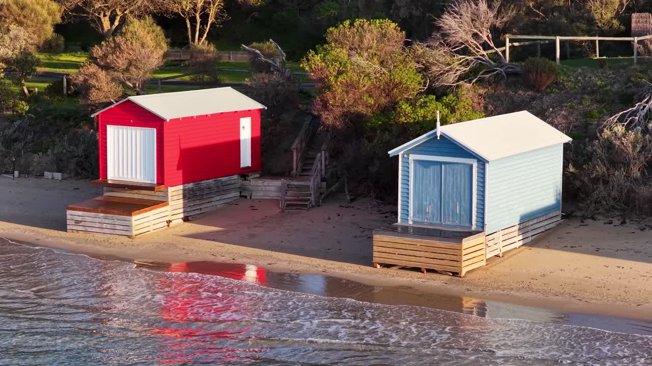 Drone pans over vibrant beach huts on sandy shoreline, golden hour sunlight, calm coastal environment