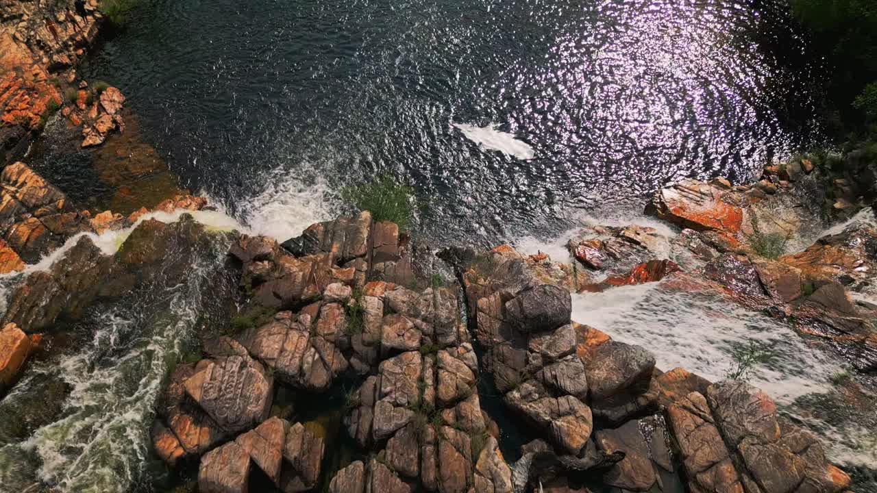 Acqua bianca che scorre giù per rocce rosse e scogliere nella cascata Cataratas dos Couros, circondata da vegetazione verde nel parco nazionale Chapada dos Veadeiros, a Goiás, Brasile, ripresa con drone con telecamera che si inclina verso il basso