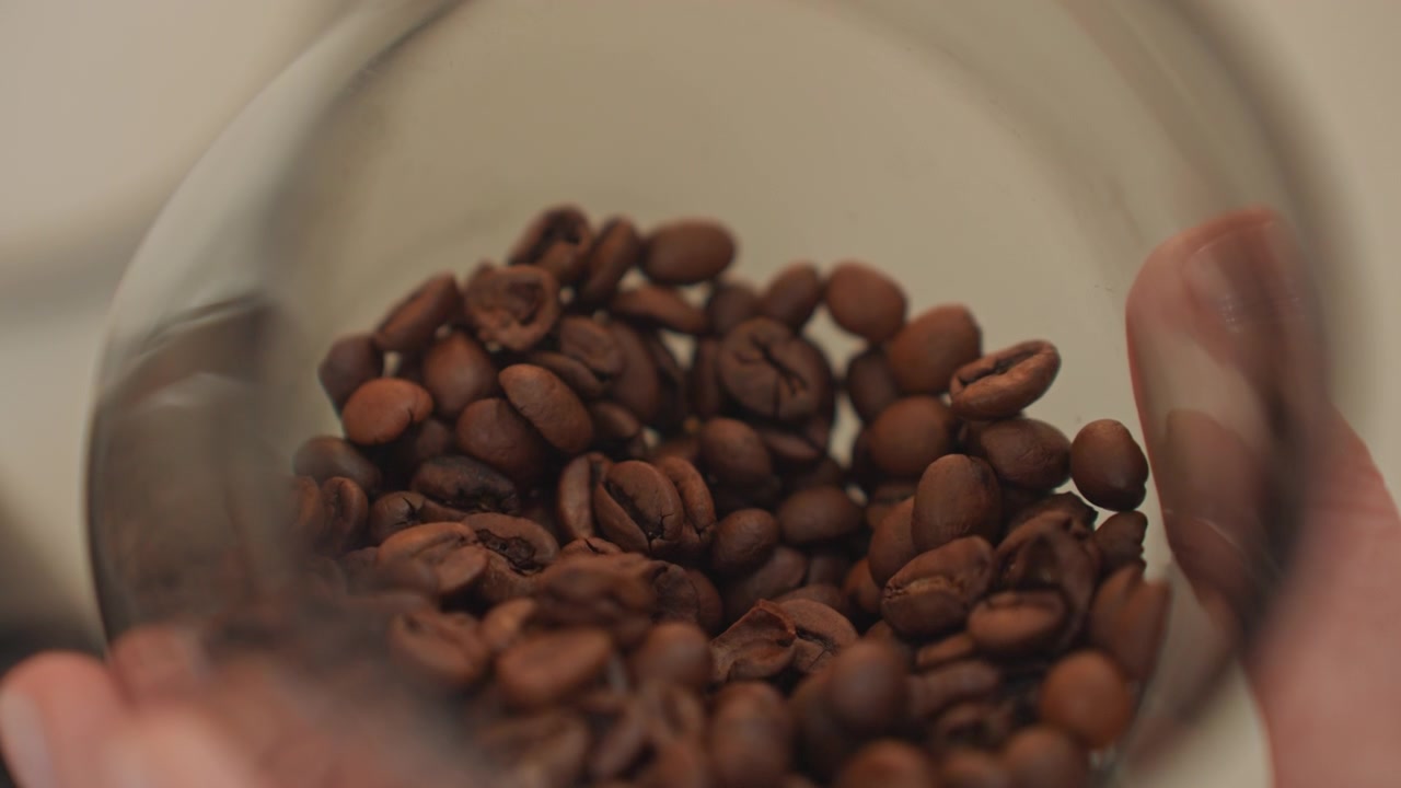 Chicchi di caffè in primo piano in un barattolo di vetro, mani che accarezzano il recipiente mentre un cucchiaio tocca brevemente i chicchi, messa a fuoco morbida e calda con tostatura marrone intenso e superficie testurizzata, atmosfera di musica acustica delicata, musicista casalingo che assapora