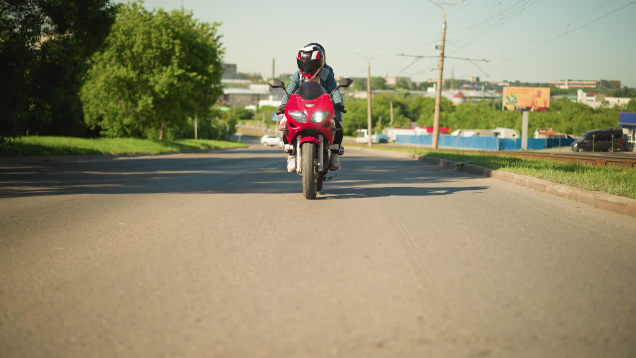 dos amigos, ambos con cascos, viajan en una bicicleta eléctrica roja a lo largo de una carretera de la ciudad, en la distancia, se ve un coche blanco haciendo un giro, con otros vehículos moviéndose en el carril opuesto