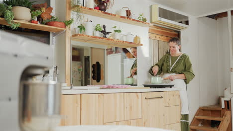 middle-aged woman in green dress and white apron stirs pot on induction cooker in cozy rustic kitchen, with large mirror reflecting background decor, plants on shelves, and pink desserts on counter