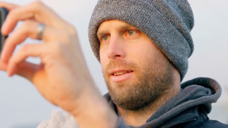 vista frontal de un hombre caucásico adulto haciendo clic en una foto con un teléfono móvil cerca de la playa 4k