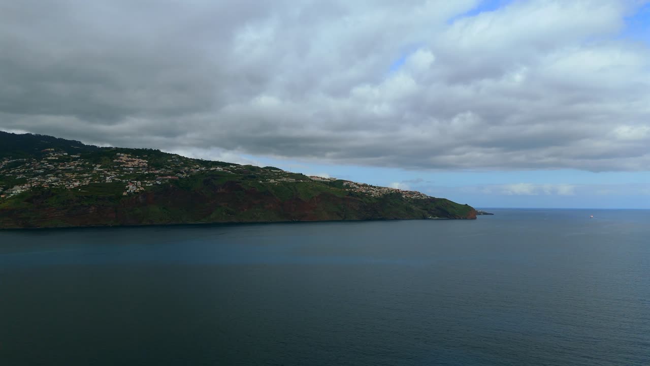 Coastal drone shot over southern Madeira showing steep green cliffs, hillside settlements and expansive views over the Atlantic under a cloudy sky