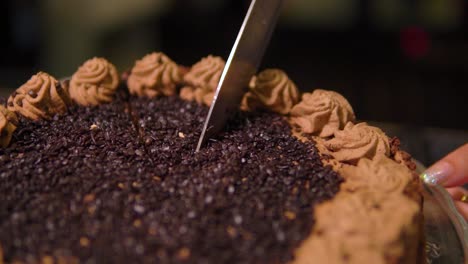 woman with shiny nails is cutting chocolate cake with decorated brown cream on top