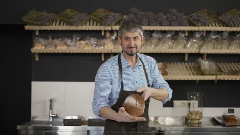 Handsome-man-in-apron-taking-fresh-bread-from-the-shelf-and-showing-it-on-camera.-Indoor,-bakery-store