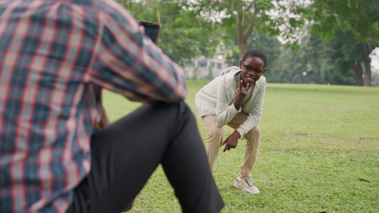 Son dancing for father in park showing energetic moves while dad records with phone, boy bending and reaching with goofy gestures, grassstained sneakers and casual outfit, laughter and spontaneous