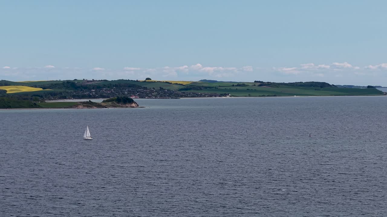 Aerial view of a white sailboat cruising on calm water with the rolling hills of Mols Bjerge in the background