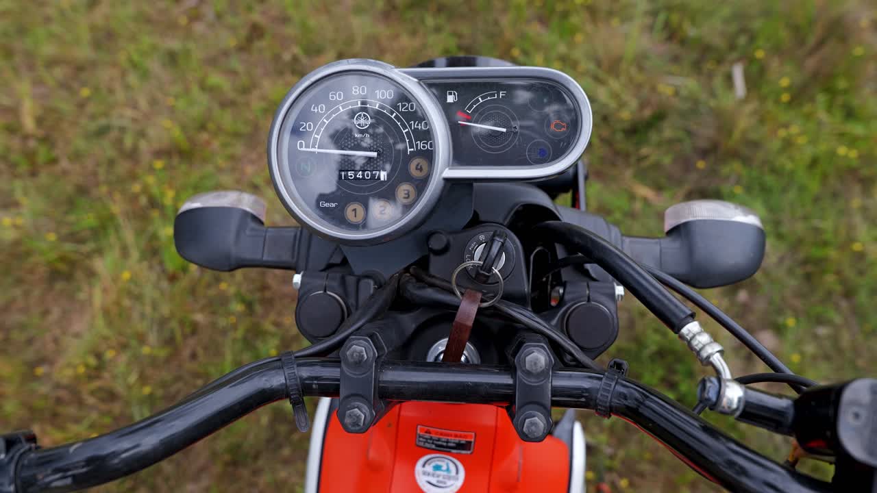 Um close-up de um b-roll mostra uma chave de ignição de motocicleta sendo ligada, com o velocímetro e o guidão visíveis no interior do Camboja
