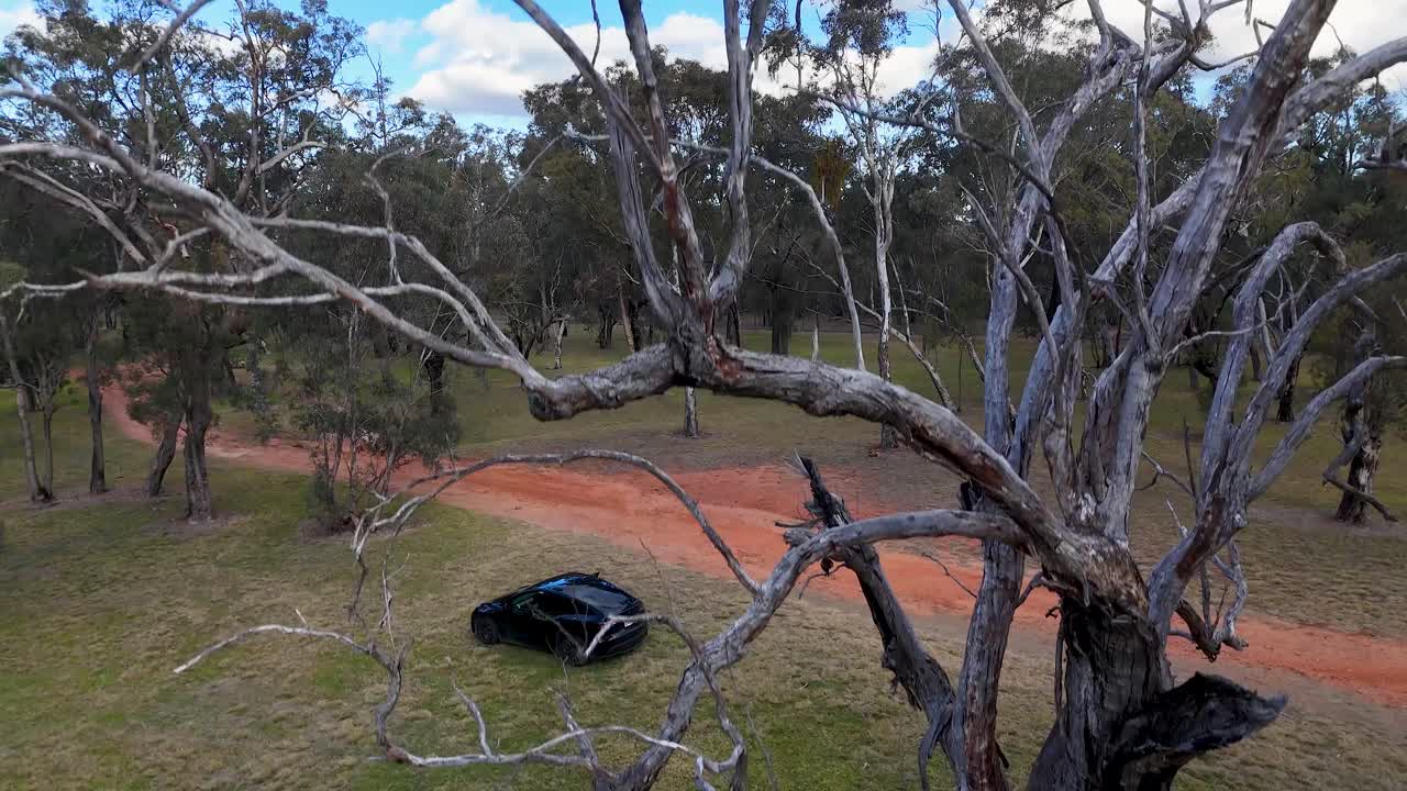 A black electric vehicle travels along a winding dirt path in a grassy, wooded park. The camera ascends past a large tree, revealing the natural landscape