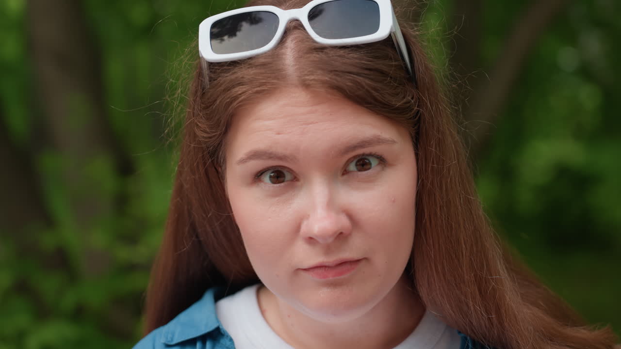 Close up of young lady sitting on wooden park bench outdoors poking thumb gently with small sharp object while focusing with serious expression, surrounded by soft green forest background