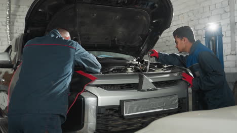 two mechanics in blue uniforms work on car engine, exchanging tools and making repairs in a professional workshop, with bright light shining from background